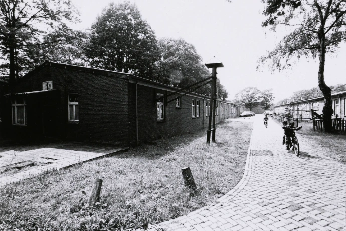 Twee kinderen fietsen langs de barakken van Kamp Vught, gefotografeerd in zwart-wit